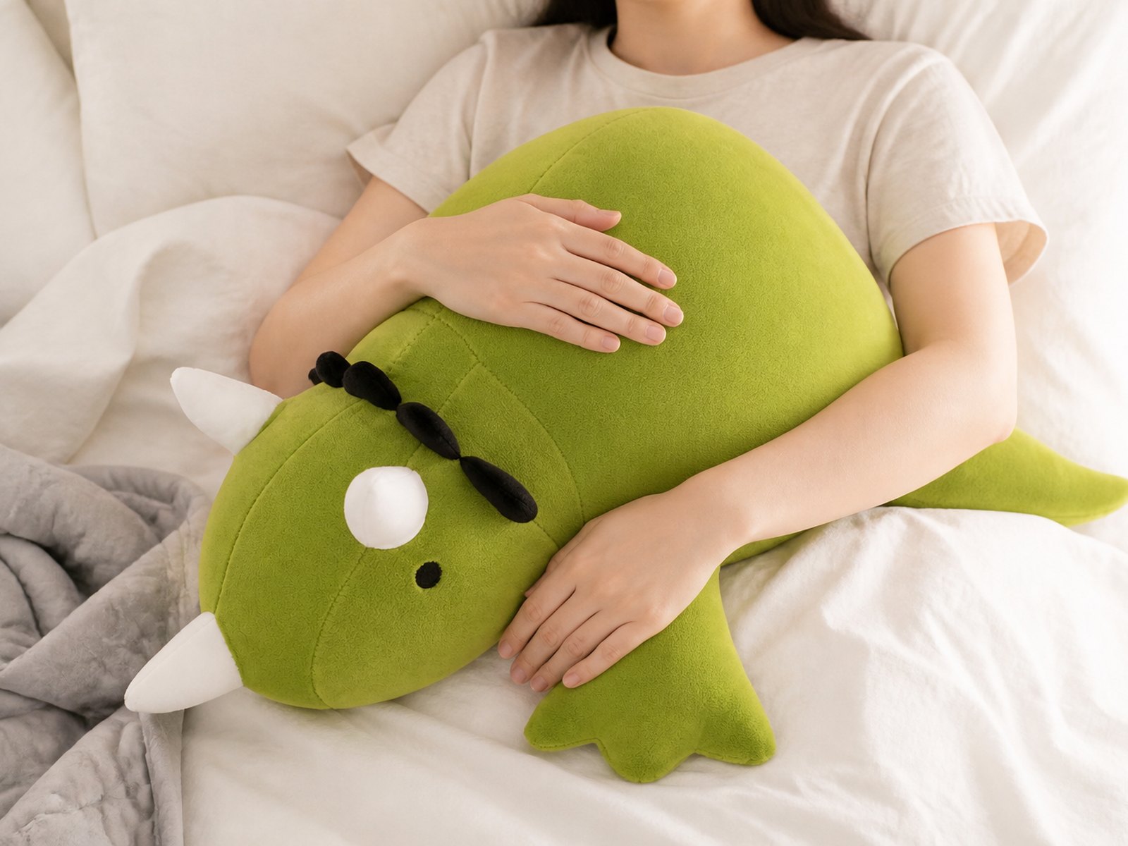 The green triceratops weighted plushie resting on someone's chest in bed, a weighted blanket pushed aside behind them