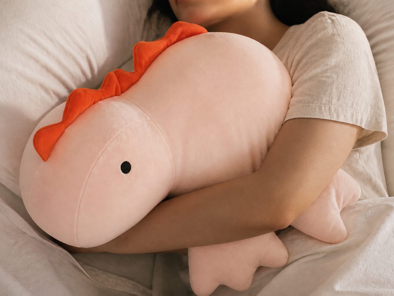 The pink dinosaur weighted plushie tucked into the crook of someone's arm in bed